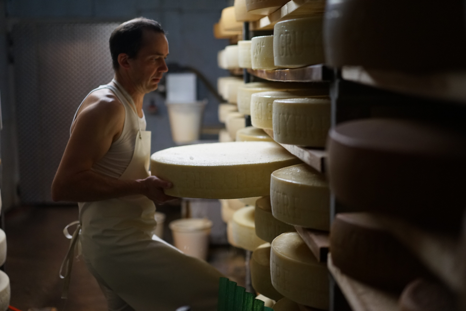 Ein Käser hebt einen großen Gruyère-Laib im Reifekeller aus dem Holzregal